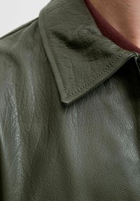 Chaqueta de cuero verde oliva con superficie texturizada, cuello con punta y detalles de costura visibles. Presenta arrugas y brillo.