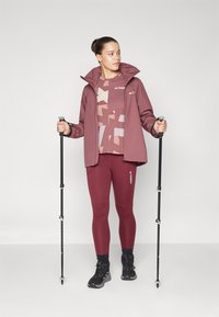 Bordo vandeniui atspari striukė, raštuotas topas ir leggingai; laiko žygių lazdas. Yra juodi batai ir elegantiškas dizainas lauko veiklai.