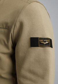 Felpa khaki con una trama liscia, maniche raglan e una toppa in tessuto nera con logo giallo e testo a contrasto.