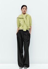 Maglione verde chiaro annodato intorno al collo, maniche corte; pantaloni neri a gamba larga; scarpe argentate; stile semplice e minimalista.