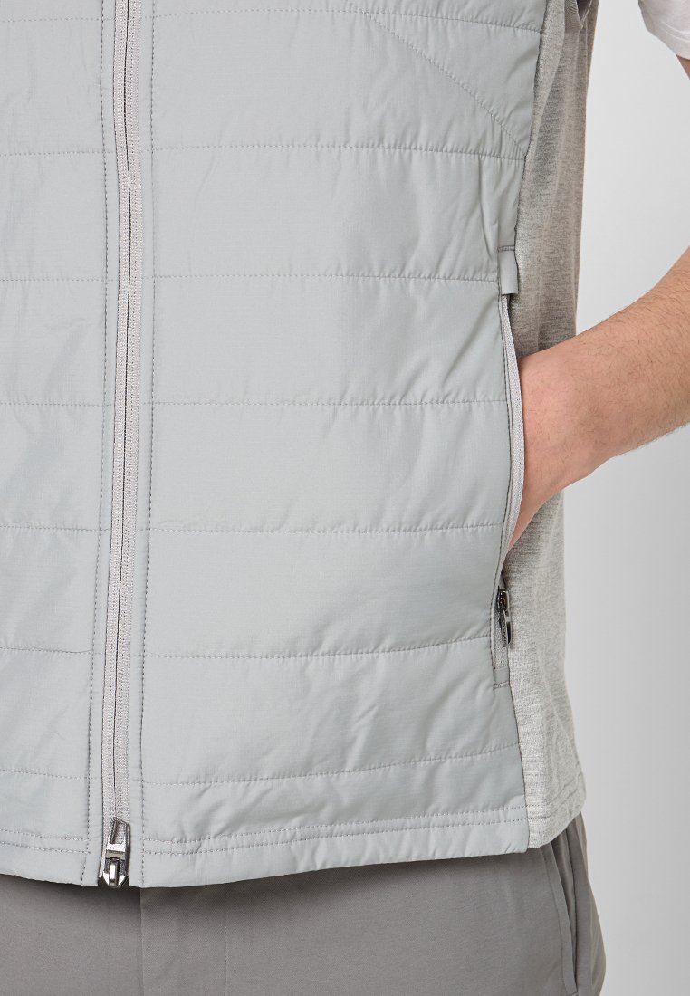 Chaleco acolchado de color gris claro con una textura suave, que presenta un zipper central, bolsillos laterales y un diseño en capas sobre una camisa gris.
