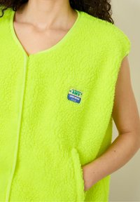 Lime green, sleeveless fleece vest with a soft texture, featuring a round neckline and a patch label on the front.