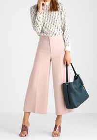 Ljusrosa byxor med vida ben, mönstrad vit blus, kombinerade med metallicfärgade slidesandaler och en mörk teal shoppingväska. Mjuka texturer, avslappnad passform.