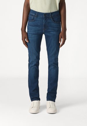 Jeans blu da uomo, slim fit, realizzati in denim, con cinque tasche e chiusura a bottoni, abbinati a sneakers bianche.