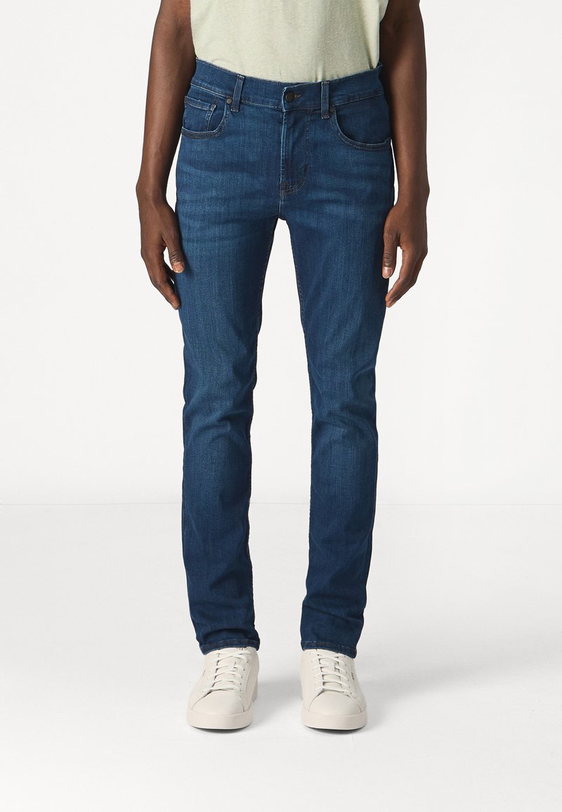 Jeans blu da uomo, slim fit, realizzati in denim, con cinque tasche e chiusura a bottoni, abbinati a sneakers bianche.
