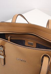 Sac à main en cuir tan de la marque Guess avec des poignées courtes, poche zippée ouverte montrant une doublure intérieure estampillée.