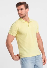 Gul polo-shirt lavet af bomuld med krave og korte ærmer. Har et lille logo på brystet og slidser i siden ved kanten.