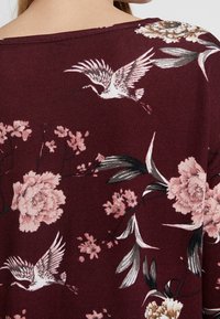 Bordeaux top met een bloemenpatroon van roze pioenrozen en kraanvogels. Zachte stof met een losse pasvorm en een brede halslijn.