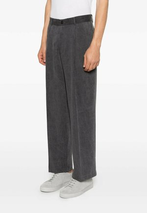 Uomo che indossa pantaloni larghi grigio scuro e scarpe da ginnastica bianche, in piedi su uno sfondo bianco semplice.