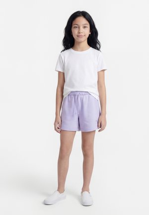 Lány áll, keze laza, fehér pólót, világoslila megkötős rövidnadrágot és fehér belebújós cipőt visel egyszerű világos háttéren.