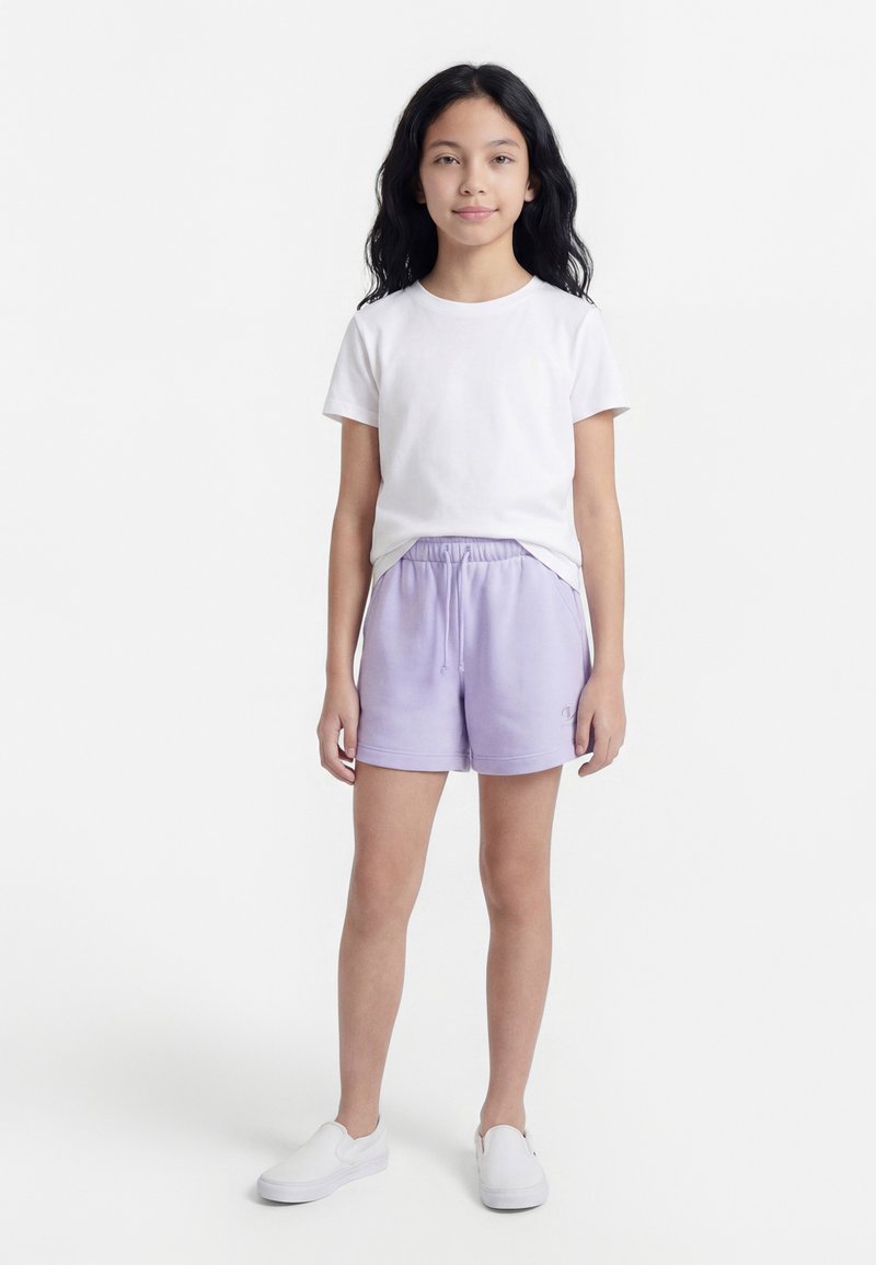 Κορίτσι στέκεται με τα χέρια χαλαρά, φορώντας λευκό μπλουζάκι, ανοιχτό μωβ σορτς με κορδόνι και λευκά παπούτσια τύπου slip-on σε απλό ανοιχτό φόντο.