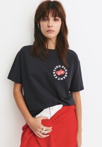 Tricou din bumbac bleumarin, cu un grafic circular alb ce include un inimă roșie și textul "Feeling Pleasant LA." Modelul poartă o fustă tricotată roșie.