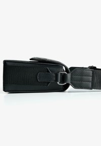 Svart texturerad rektangulär crossbody-väska med justerbar rem och klaffstängning, som ligger på en vit yta.