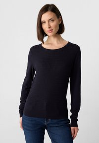 Frau mit braunem Bob-Haarschnitt, die einen strukturierten navyblauen Pullover und blaue Jeans trägt, steht vor einem schlichten hellen Hintergrund.