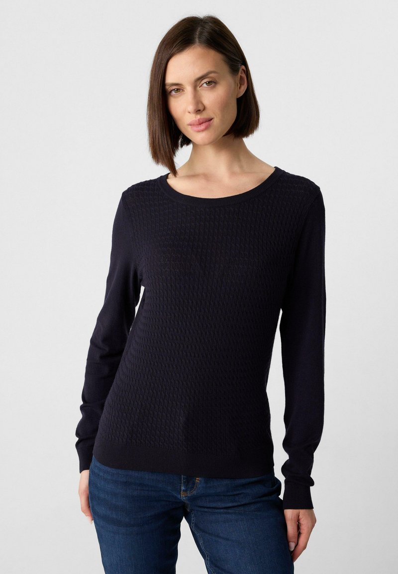 Frau mit braunem Bob-Haarschnitt, die einen strukturierten navyblauen Pullover und blaue Jeans trägt, steht vor einem schlichten hellen Hintergrund.