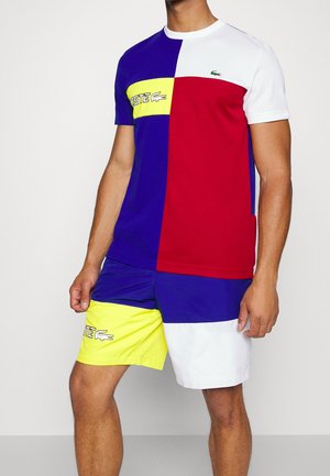 Hombre vistiendo una camiseta y pantalones cortos bloqueados por colores en azul, rojo, blanco y amarillo con detalles de logotipo en el pecho y el muslo izquierdo.