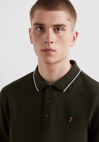 Dunkelgrünes Poloshirt mit einem kontrastierenden kragen in Weiß und Burgund gestreift, mit einem kleinen gestickten Logo auf der linken Brust.