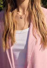 Henkilö, jolla on pitkät vaaleat hiukset, käyttää kerroksellisia kultaketjuja ja vaaleanpunaista bleiseriä valkoisen ribbipaitansa päällä auringonvalossa.