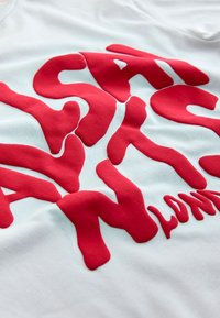 Hvid T-shirt med markante, hævede røde bogstaver, der danner ordet "SANTS" i et flydende, abstrakt design. Glat tekstur.