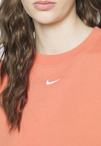 Młoda osoba z długimi falującymi brązowymi włosami, ubrana w koralowy t-shirt Nike z małym białym logo Swoosh przy kołnierzu.