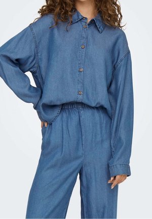 Kobieta ubrana w luźną niebieską jeansową koszulę na guziki oraz dopasowane spodnie z elastycznym pasem, stojąca z jedną ręką na biodrze.