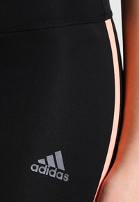 Černé sportovní legíny s úzkým střihem, měkkou texturou a růžovým bočním pruhováním, s šedým logem Adidas blízko spodní části.