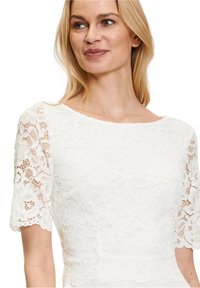 White lace top featuring short sleeves, floral patterns, and a boat neckline, with a textured finish and scalloped hem details.