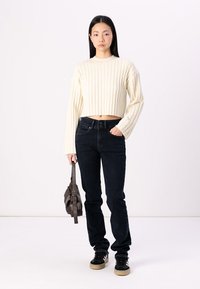 Cream ribbed cropped sweater, dark blue jeans, black sneakers with tan soles, holding a brown handbag. Plain white background.