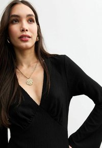 Robe noire côtelée à manches longues, dotée d'un décolleté en V profond et d'un collier en pendentif texturé doré.