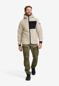 Fleecejacka i beige med en svart bröstficka och dragkedja, i kombination med gröna cargobyxor med dragkedjefickor och en logotyp-patch.