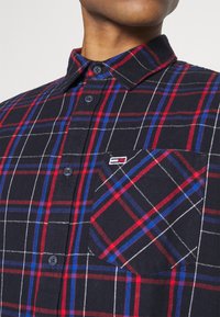 Koszula w kratę w kolorze granatowym, czerwonym i niebieskim z białymi akcentami. Posiada kołnierzyk zapinany na guziki, kieszonkę z przodu oraz teksturowany materiał.