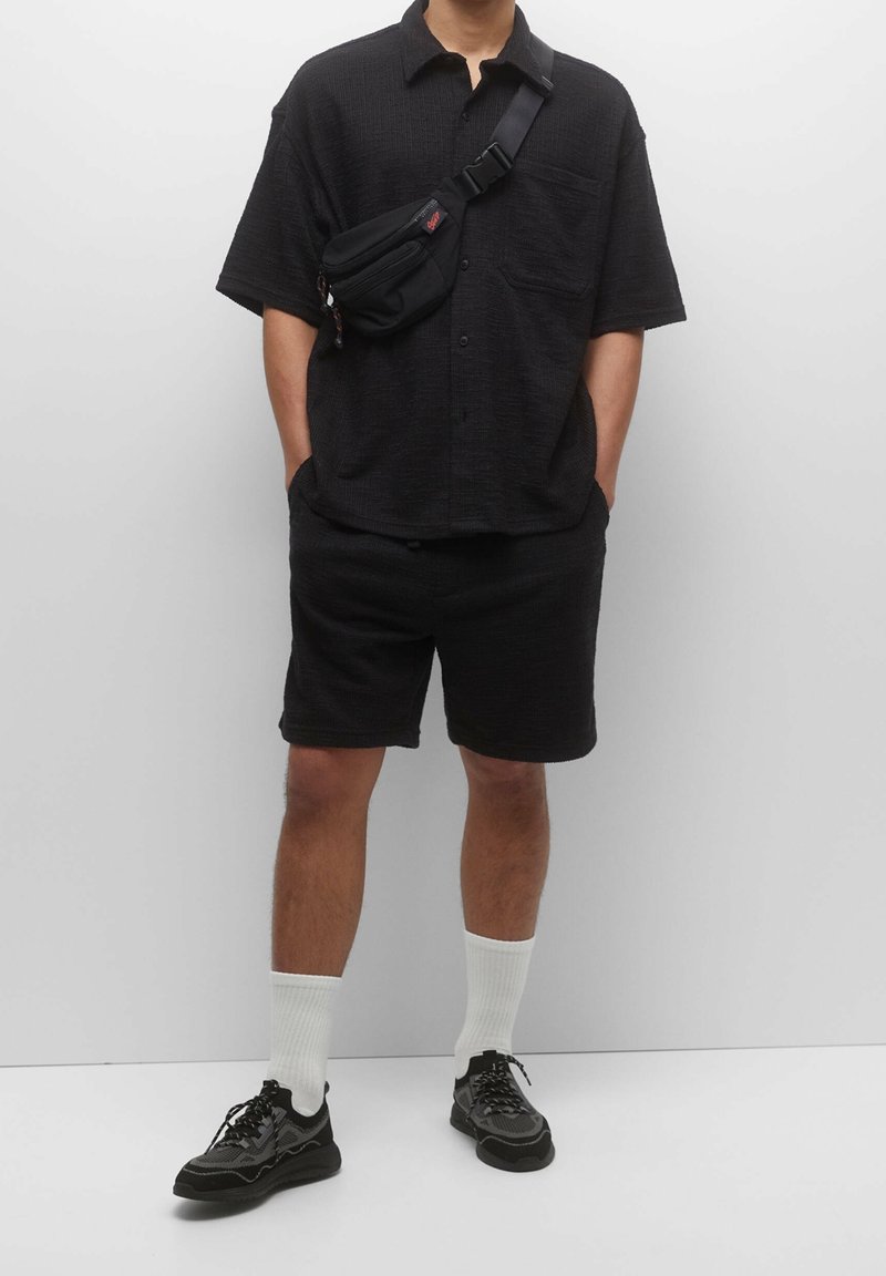 Camisa negra de manga corta con un patrón texturizado, pantalones cortos negros, zapatillas negras y calcetines blancos hasta el tobillo. Pequeño bolso negro de hombro con detalle de cremallera.