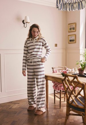 Vrouw in een gestreept shirt met lange mouwen en broek, slippers aan, staat bij een houten eettafel in een zacht verlichte kamer met ingelijste foto's en planten.