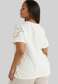 Camiseta blanca de manga corta, con un intrincado detalle de recortes en los hombros y un ajuste holgado. Material texturizado y ligero.