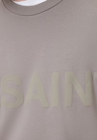 Svetlo šedý sveter s hladkou textúrou. Má výrazné, zvýšené písmo, ktoré hovorí "SAINT" v o niečo tmavšom odtieni. Okrúhly výstrih.