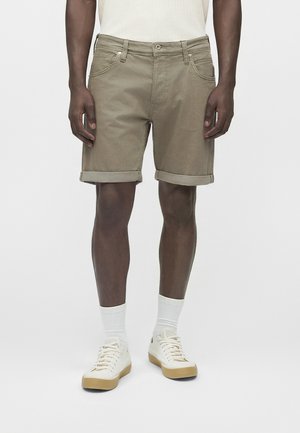 Mann trägt beige Shorts mit umgeschlagenem Saum, weiße Knöchelsocken und weiße Sneaker, steht vor einem schlichten, hellen Hintergrund.