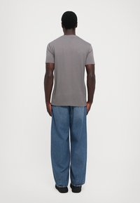 Mann som står vendt bort fra kameraet iført en grå kortermet skjorte, løse blå jeans og svarte sko mot en enkel hvit bakgrunn.