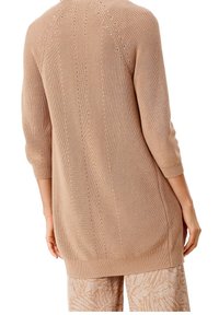 Ljusbrun stickad tröja med ett texturerat mönster, som har ärmar i tre fjärdedelar, ribbad mudd och nederkant. Längden slutar vid höfterna.