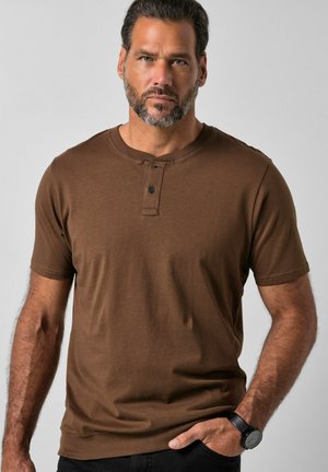 Bärtiger Mann mit kurzem dunklem Haar, der ein braunes kurzärmeliges Henley-Shirt, eine schwarze Uhr und schwarze Hose trägt, steht vor einem einfarbigen Hintergrund.