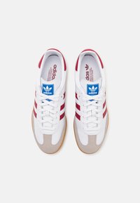 White athletic shoes featuring red stripes, soft suede accents, rubber sole, and a blue logo tag on the tongue. Classic design with lace closure.