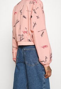 Růžová bomber bunda s elastickými manžetami zdobená černými a červenými graffiti nápisy a grafikou, doplněná o modré džíny s širokými nohavicemi.