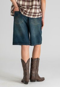 Shorts en denim bleu foncé, longueur mi-cuisse, avec poches arrière ornées de coutures décoratives ; associés à des bottes de cow-boy marron avec des motifs complexes.