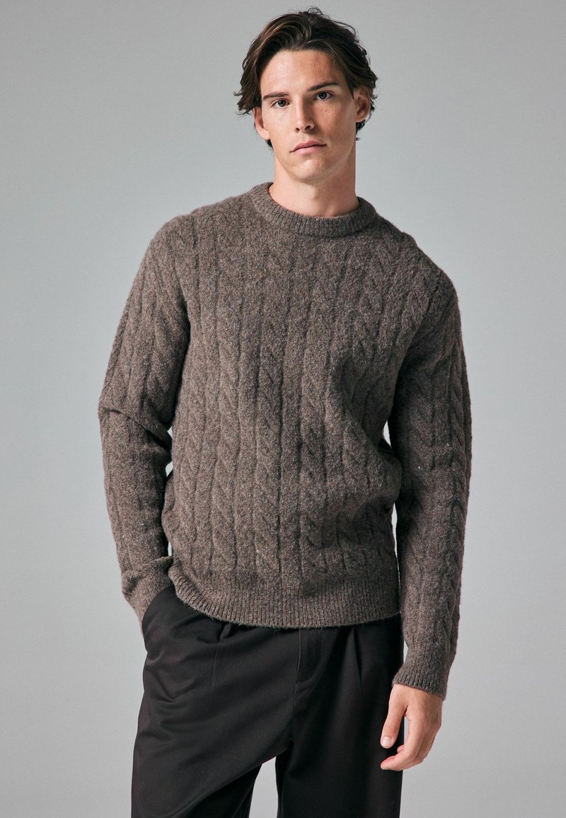 Pulover maro din tricot cu model împletit, cu guler rotund și mâneci lungi, având mansete și tiv cu dungi. Purtați peste pantaloni închiși la culoare.