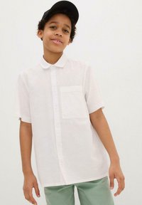 Camicetta bianca a maniche corte con colletto e una tasca sul petto, indossata con pantaloni verde chiaro. Tessuto morbido, design casual.