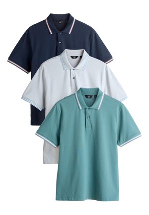 Mand med langt flet hår iført lysegrå poloshirt, mørke jeans med lige ben og brune snørebukser, der står op ad en hvid væg.