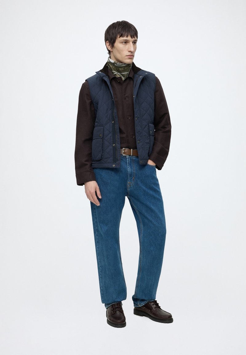 Ung mand iført en mørkebrun skjorte, blå vatteret vest, blå jeans, mønstret grøn tørklæde og mørkebrune sko står med en hånd i lommen.