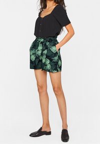 Svart kortärmad blus med knappar, tillsammans med svarta shorts med gröna bladmönster och sidofickor. Svarta slip-on skor.