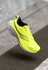 Helle gelbe Laufschuhe mit grauer Sohle in der Luft über einer gepflasterten Straße mit weißen und grauen Linien.