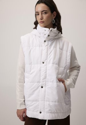 Žena v bielej prešívanej bezrukávovej bunde so zapínaním na patentky cez béžový dlhý rukáv svetra, s jednou rukou vrecku, neutrálne pozadie.