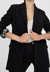 Czarna marynarka o strukturalnym kroju, z klapą z wycięciami, dwoma przednimi kieszeniami oraz wzorzystymi mankietami na rękawach. W zestawie z jasnym topem.